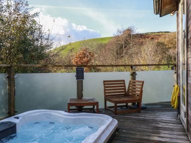 An outdoor wooden deck with a hot tub wooden chairs and table overlooking trees and hills at 3 Hedgerows in Lanreath