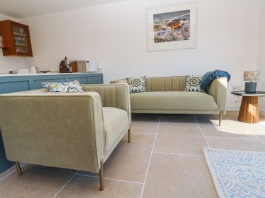 A living room with two sofas a side table with a lamp and a kitchen counter with appliances at Little Victory in Pendeen