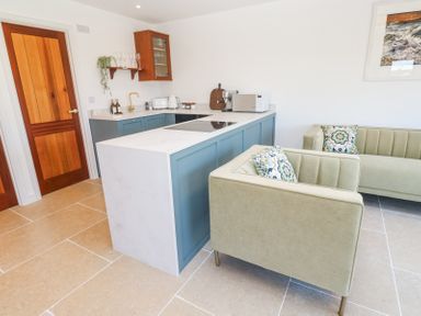 A kitchen area with blue cabinets a white countertop a wooden door beige tiled floor and green upholstered armchairs at Little Victory in Pendeen