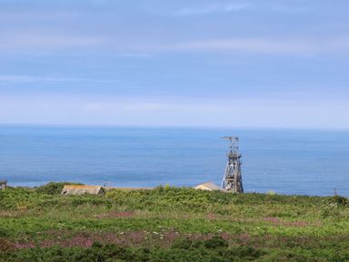 A coastal view with a metal structure and buildings behind green shrubbery and flowers at Little Victory in Pendeen