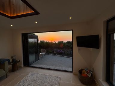 A room with a rug on the floor and a wall-mounted TV next to an open large door showing an outdoor patio at sunset at Little Victory in Pendeen