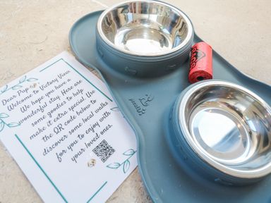 A pet feeding station with two stainless steel bowls a roll of red dog waste bags and a welcome note for dogs at Little Victory in Pendeen