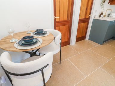 A small dining area with a round wooden table set for two and white cushioned chairs near wooden doors and a kitchen counter at Little Victory in Pendeen