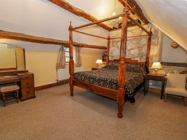 A bedroom with a wooden four-poster bed bedside tables with lamps a dressing table with a stool an armchair and exposed wooden beams at Ysgubor in Dinas Mawddwy near Dolgellau