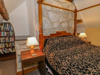 A bedroom with a wooden four-poster bed covered with a patterned bedspread and two bedside tables with lamps near a stone wall and a bookshelf at Ysgubor in Dinas Mawddwy near Dolgellau