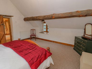 A bedroom with a wooden bed with red bedding a wooden chair a chest of drawers with a mirror and a window under a wooden beam at Ysgubor in Dinas Mawddwy near Dolgellau