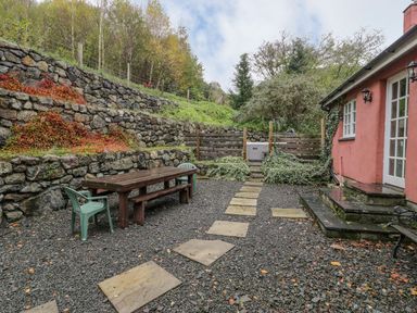 An outdoor patio area with a wooden table and plastic chairs on gravel surrounded by stone retaining walls and a pink building at Ysgubor in Dinas Mawddwy near Dolgellau