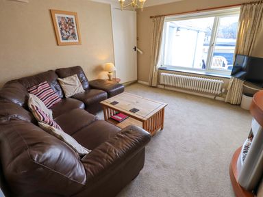 A living room with a brown leather sofa a wooden coffee table a floor lamp a wall painting a large window with curtains and a television at Lakeland View Lodge in Keswick