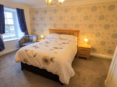 A bedroom with a double bed between two bedside tables with lamps a window with blue curtains and an armchair at Lakeland View Lodge in Keswick