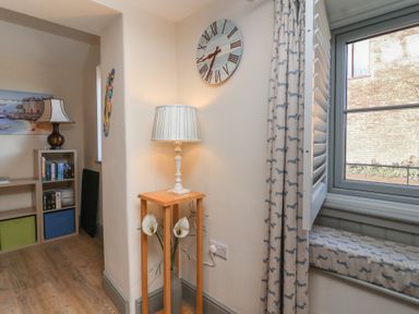 A corner with a wooden side table holding a lamp and vase near a window with patterned curtains and a shelf with books and a lamp at Curlew Cottage in North Curry near Taunton