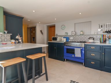 A kitchen with blue cabinets a blue oven a white countertop stools and patterned tile backsplash at Curlew Cottage in North Curry near Taunton