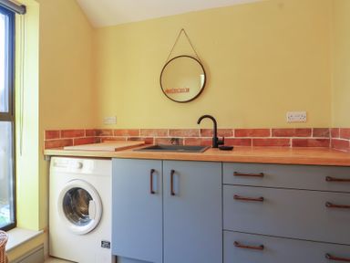 A kitchen area with blue cabinets a washing machine a black faucet a round mirror and brick backsplash at Curlew Cottage in North Curry near Taunton