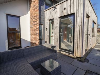 An outdoor patio area with gray woven sectional sofa and a small glass-top table next to a wooden and brick exterior wall at Curlew Cottage in North Curry near Taunton