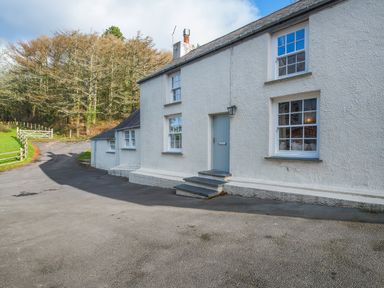 The exterior of a white house with multiple windows and a light blue door next to a paved driveway and a wooded area at Leeward Cottage in Cosheston near Pembroke Dock