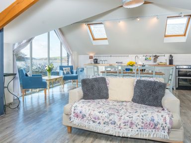 An open living area with a sofa and two blue chairs near large windows overlooking water at Leeward Cottage in Cosheston near Pembroke Dock