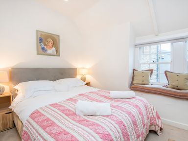 A bedroom with a bed covered by a pink patterned quilt and white rolled towels two bedside tables with lamps a window seat with cushions and a painting above the bed at Leeward Cottage in Cosheston near Pembroke Dock