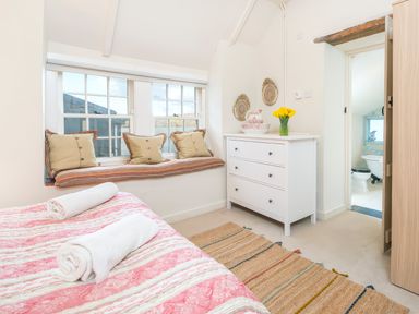 A bedroom with a bed covered in a pink and white striped quilt with two rolled white towels a window seat with cushions a white dresser with flowers and bowls and a glimpse of a bathroom at Leeward Cottage in Cosheston near Pembroke Dock