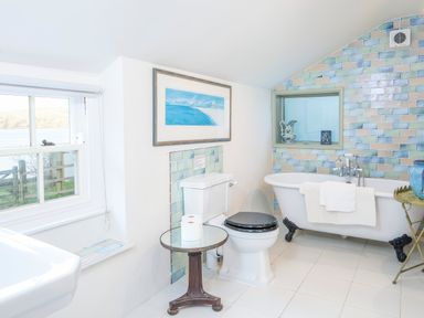 A bathroom with a toilet black seat a clawfoot bathtub with towels a small round table with toilet paper a window and decorative tiled wall at Leeward Cottage in Cosheston near Pembroke Dock
