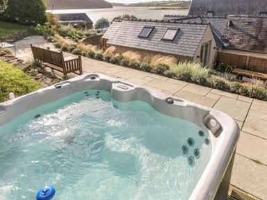 A hot tub on a patio near a bench and plants with houses and a body of water in the background at Leeward Cottage in Cosheston near Pembroke Dock