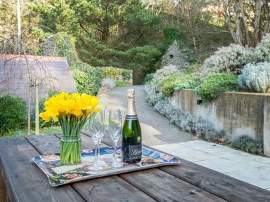 A wooden outdoor table with a tray holding a bottle of champagne a vase of yellow flowers and two empty glasses at Leeward Cottage in Cosheston near Pembroke Dock