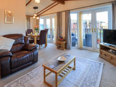 A living room with a brown leather sofa coffee table dining table with chairs and large windows at Lavender Lodge South Lakeland Leisure Village