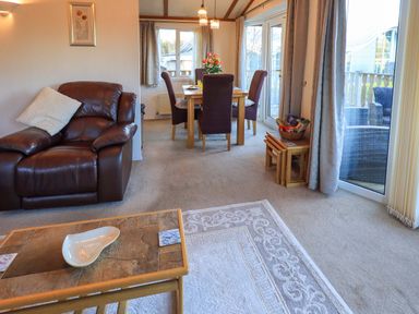 A living room with a brown leather armchair a wooden coffee table with a ceramic dish a dining table with four purple chairs and flowers on it at Lavender Lodge in South Lakeland Leisure Village