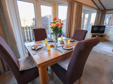A dining area with a wooden table set for four with croissants drinks and a vase of flowers adjacent to large windows in a living room at Lavender Lodge in South Lakeland Leisure Village