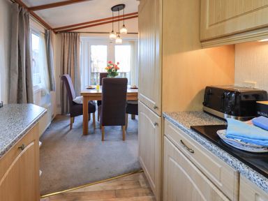 A dining area with a wooden table and four chairs next to glass doors and a kitchen counter with a toaster and folded towels at Lavender Lodge in South Lakeland Leisure Village