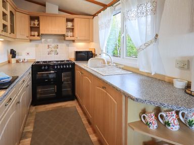 A kitchen with wooden cabinets a black oven a sink under a window and decorative pitcher mugs on a shelf at Lavender Lodge in South Lakeland Leisure Village