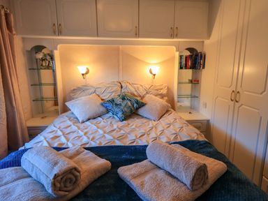 A bedroom with a bed with cushions folded towels on a blanket overhead cabinets and a bookshelf at Lavender Lodge in South Lakeland Leisure Village