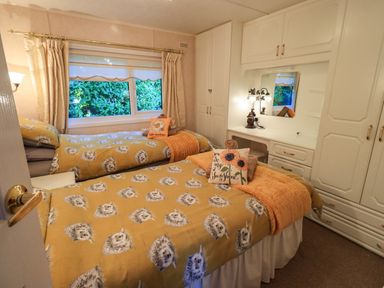 A bedroom with two single beds pillows and blankets a window with curtains and built in wardrobes and dressing table at Lavender Lodge in South Lakeland Leisure Village