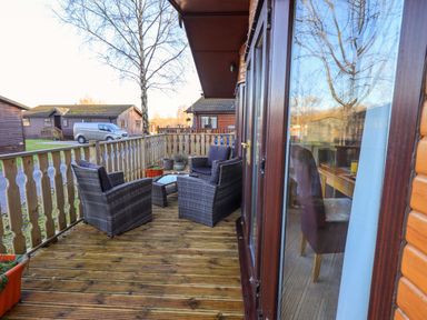 A wooden deck with wicker chairs and a table outside wooden cabins with a van in the background at Lavender Lodge in South Lakeland Leisure Village