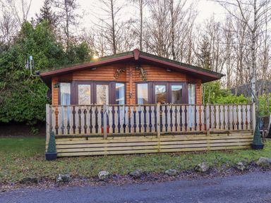 A wooden cabin with a fenced porch and glass doors surrounded by trees at Lavender Lodge in South Lakeland Leisure Village