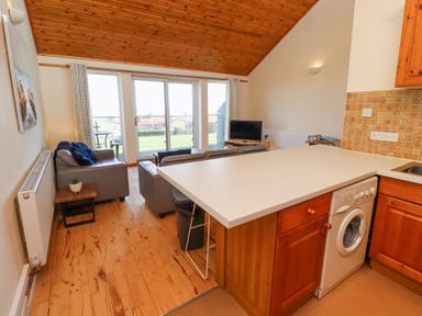 A living room with sofas a TV and glass doors leading outside next to a kitchen counter with wooden cabinets at Waters Edge in Embleton