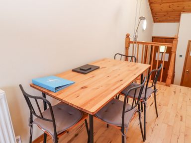 A wooden dining table with four chairs with cushions and a blue folder on the table in a room with wooden floor and railing at Waters Edge in Embleton