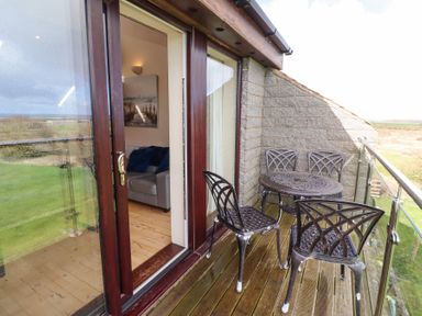 A balcony with four metal chairs and a round metal table overlooking a grassy area and a room with a sofa visible through sliding glass doors at Waters Edge in Embleton