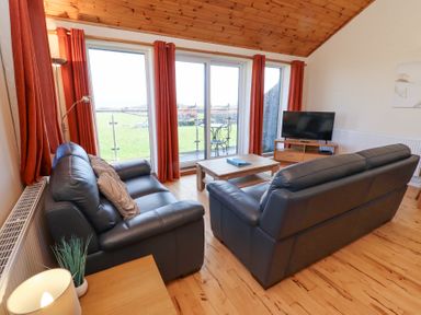 A living room with two black leather sofas a wooden coffee table and a television near large windows with orange curtains at Dunstanburgh View in Embleton