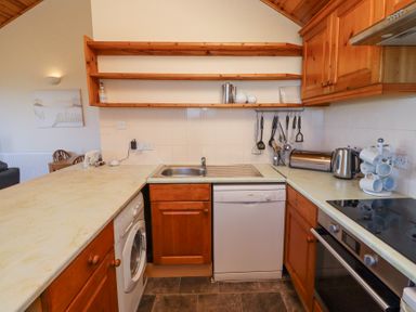 A kitchen with wooden cabinets, a washing machine, dishwasher, sink, stove, utensils hanging on the wall, and a countertop with a kettle and mugs at Dunstanburgh View in Embleton