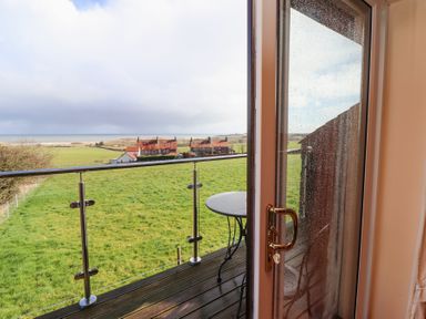 A balcony with a round table overlooking green fields and houses at Dunstanburgh View in Embleton