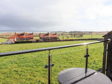 A balcony with glass railing overlooking green fields and houses with red roofs at Dunstanburgh View in Embleton