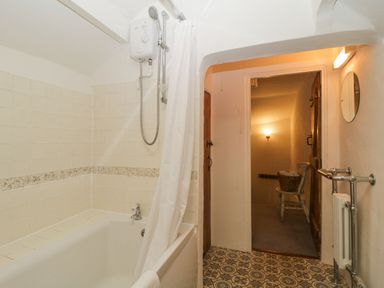 A bathroom with a bathtub shower and curtain and a chair with a basket of towels in the adjacent room at 3 Cumberland Cottages in Ringmore