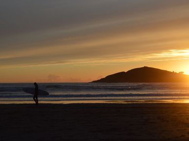 A surfer walking on a beach at sunset with an island in the background at 3 Cumberland Cottages in Ringmore