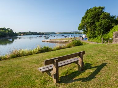 A wooden bench on grass overlooking a river with boats and trees in the background at 3 Cumberland Cottages in Ringmore