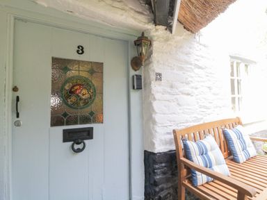 A wooden bench with striped cushions next to a white stone wall and a light blue door with stained glass at 3 Cumberland Cottages in Ringmore