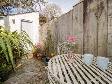 A small outdoor patio with stone flooring potted plants a white metal table with two cups and a vase of pink flowers a wooden fence and a white shed door at 3 Cumberland Cottages Ringmore