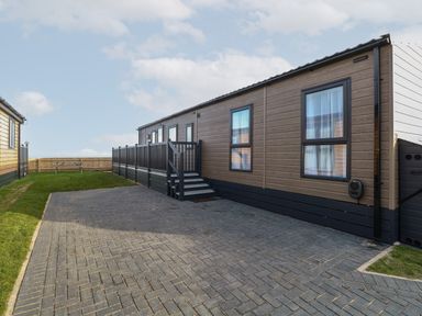 A driveway with a paved parking area next to a single story home with wooden siding and a fenced deck at Dorchester in Broomholm near Mundesley