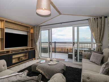 A living room with sofas a TV unit and sliding glass doors opening to a balcony with ocean view at Dorchester in Broomholm near Mundesley