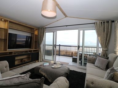 A living room with sofas a TV cabinet and sliding glass doors opening to a balcony overlooking the sea at Dorchester in Broomholm near Mundesley