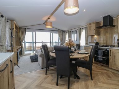 A dining table with four chairs in an open plan kitchen and living area with a sofa and sliding glass doors at Dorchester in Broomholm near Mundesley
