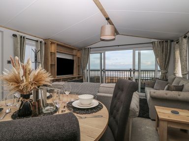 A living room with a dining table set near a large window overlooking the sea with sofas and a wooden TV unit at Dorchester in Broomholm near Mundesley